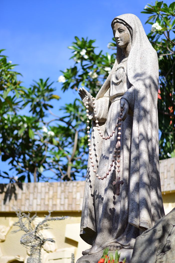 Outdoor stone statue of Virgin Mary holding a rosary, surrounded by lush greenery.
