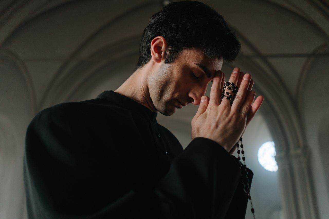 A priest in a black gown solemnly prays with rosary beads in a serene church setting.
